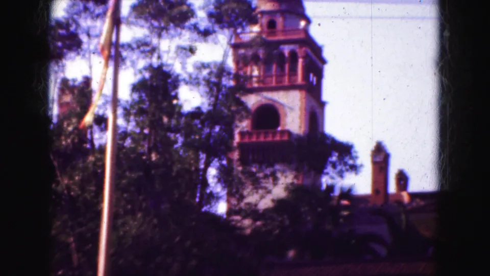 1975 Florida frame showing a bell tower with arched openings above trees, red tiled rooflines, and a bright clear sky.
