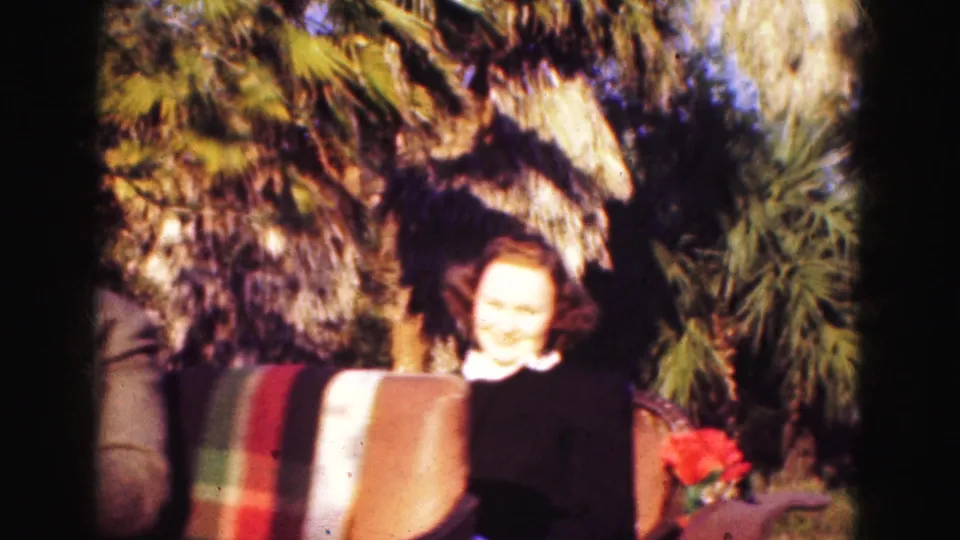 1975 Florida home-movie frame of a smiling woman passenger in a carriage, with palms, upholstery, and red flowers visible nearby.