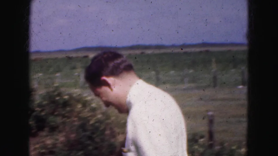 A man walks through the Everglades farm scene in a white shirt, with pasture, brush, fence line, and distant hills or tree line behind him.