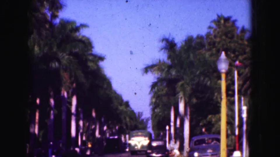 1975 Florida home-movie frame of older cars and a bus-like vehicle on a sunny palm-lined street.