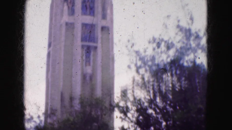 A tall pale building appears beyond trees in grainy 1975 8mm footage, linking the wooded setting with nearby urban architecture.