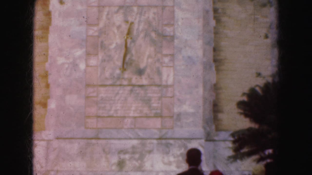 Stone monument wall with carved panel, foliage, and small visitor silhouettes.