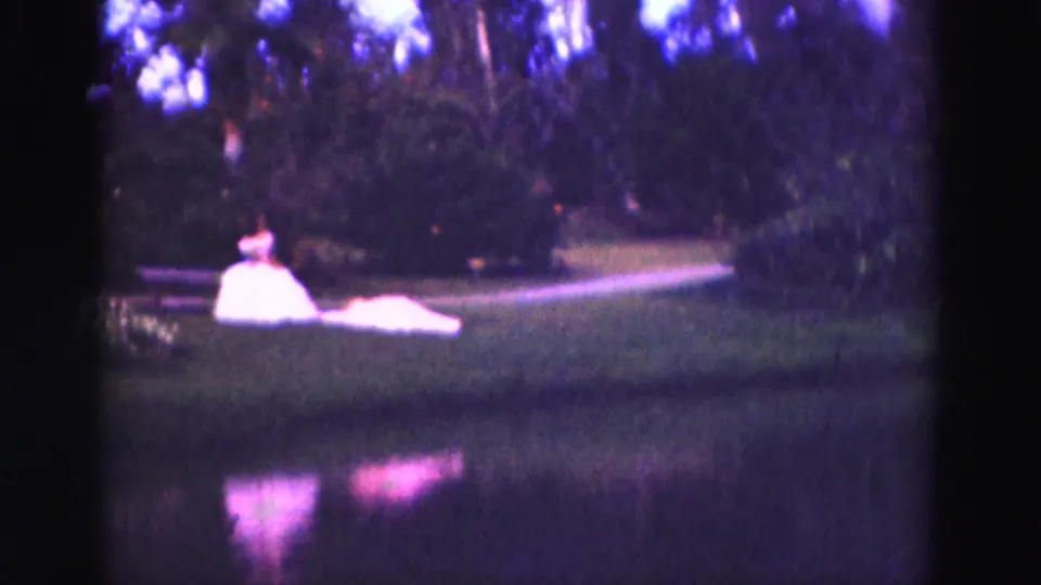 A woman in a white gown remains seated near the canal, framed by grass, water and trees in 1975 8mm footage.