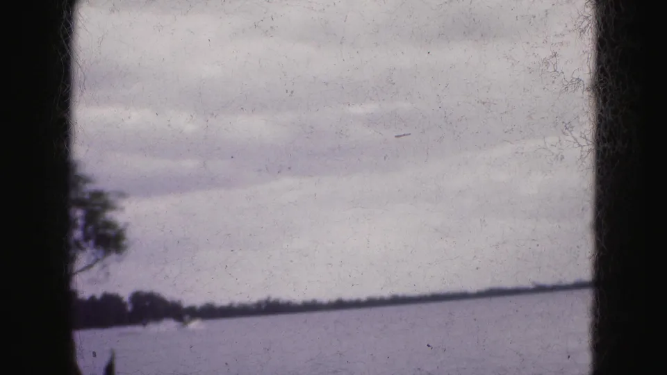 1975 Florida Everglades home-movie frame showing a wide lake surface, low shoreline, trees, and dark film gate edges.