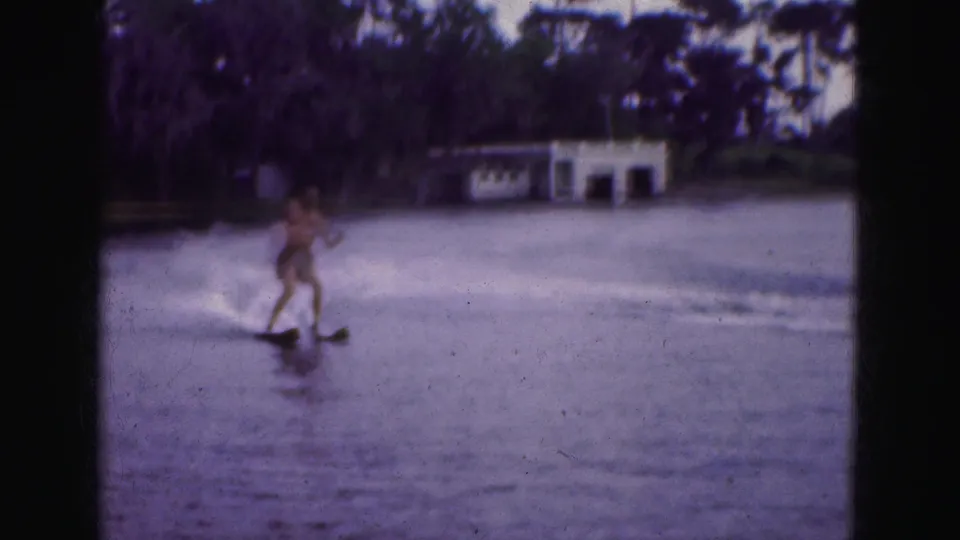 A distant water skier appears on a 1975 Florida Everglades lake, passing a wooded shoreline and small white lakeside structure.