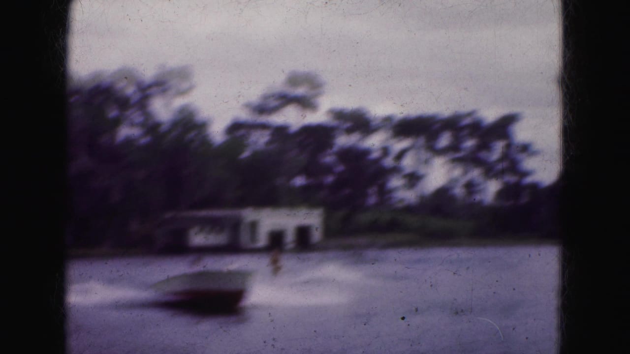 Blurred motorboat crossing a lake near a white boathouse and tree-lined shore.