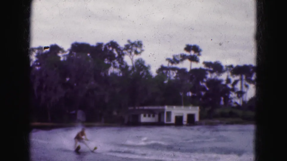 1975 Florida Everglades frame showing a man water skiing across a lake near a white boathouse and wooded shoreline.