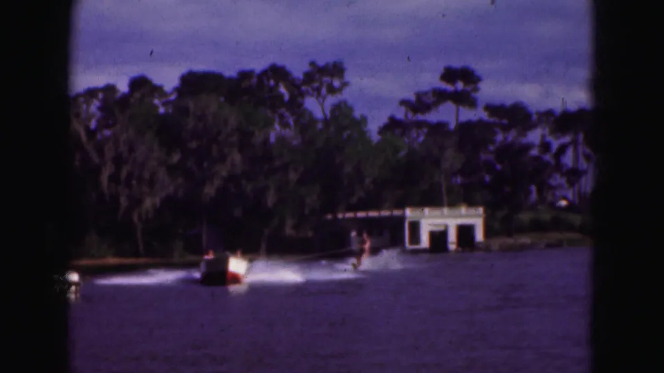 A home-movie frame shows the boat and skier crossing the lake, with dense greenery and a white boathouse in the background.
