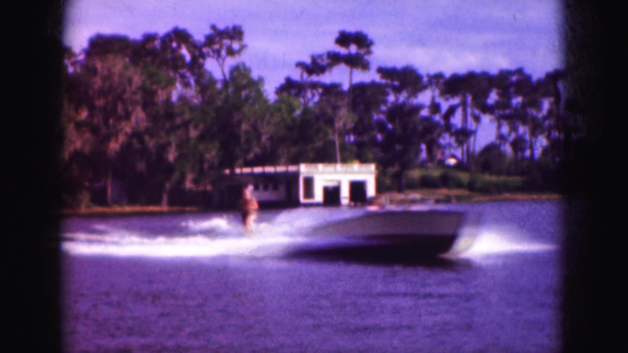 1975 Florida 8mm home-movie frame showing a water skier being pulled behind a motorboat on a lake, with a white boathouse and dense shoreline trees in the background.