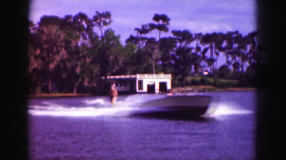 1975 Florida 8mm home-movie frame showing a water skier being pulled behind a motorboat on a lake, with a white boathouse and dense shoreline trees in the background.