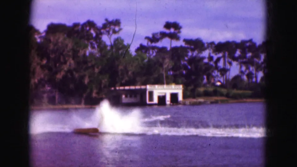 1975 Florida 8mm home-movie frame with a water skier falling into a large splash while the tow boat moves across the lake near a white boathouse.