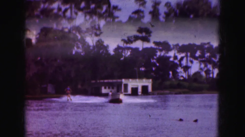 1975 Florida 8mm home-movie frame showing the water skier and tow boat farther from the camera, with lake water, shoreline trees, and a white boathouse behind them.
