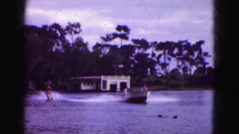 1975 Florida 8mm home-movie frame showing the skier back on the water as the motorboat speeds across the lake in front of a white boathouse and subtropical tree line.