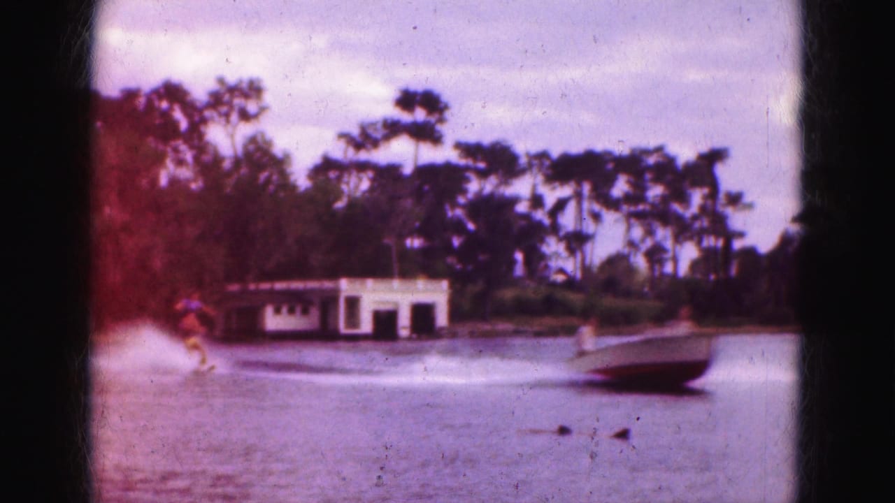 Water skier follows a speeding motorboat past a small white boathouse on a lake.