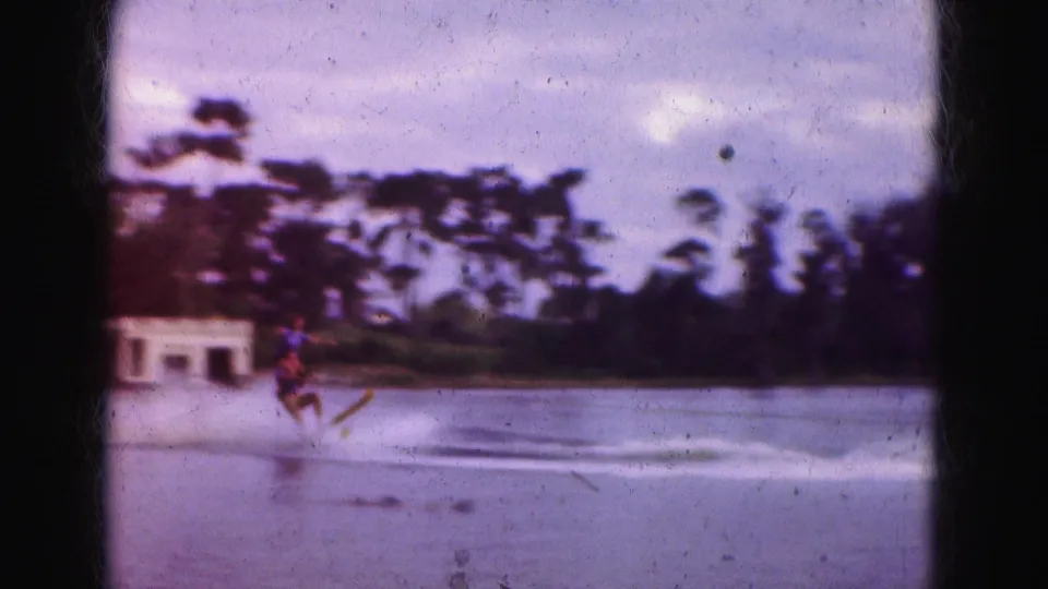 1975 Florida Everglades 8mm frame of a skier crossing the lake behind a motorboat with palms and a small boathouse beyond.