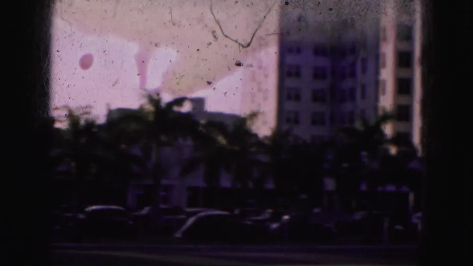 1975 Florida home-movie frame showing a shadowed palm-lined road with cars and tall buildings beyond the film gate.