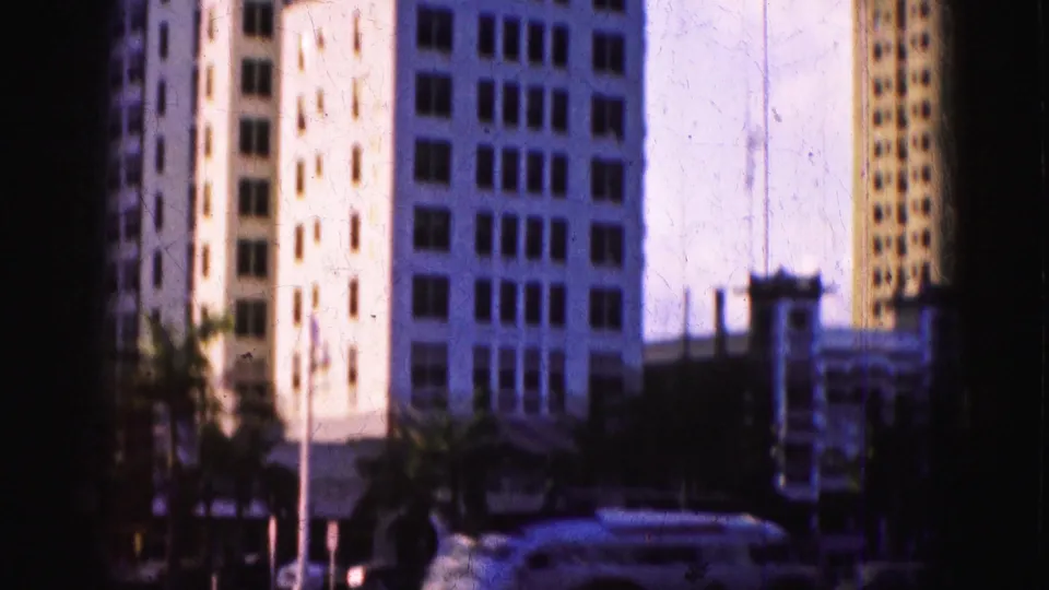 1975 Florida street frame with a white station wagon, palm trees, and tall buildings seen through soft 8mm exposure.