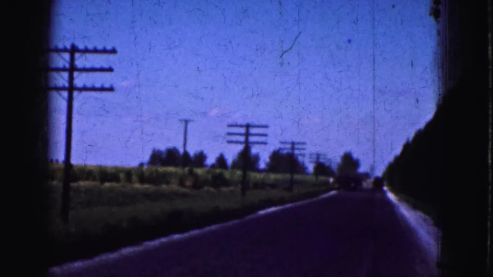 Passenger-view 1975 8mm footage of a straight Everglades-area road with utility poles, fields, and distant trees.