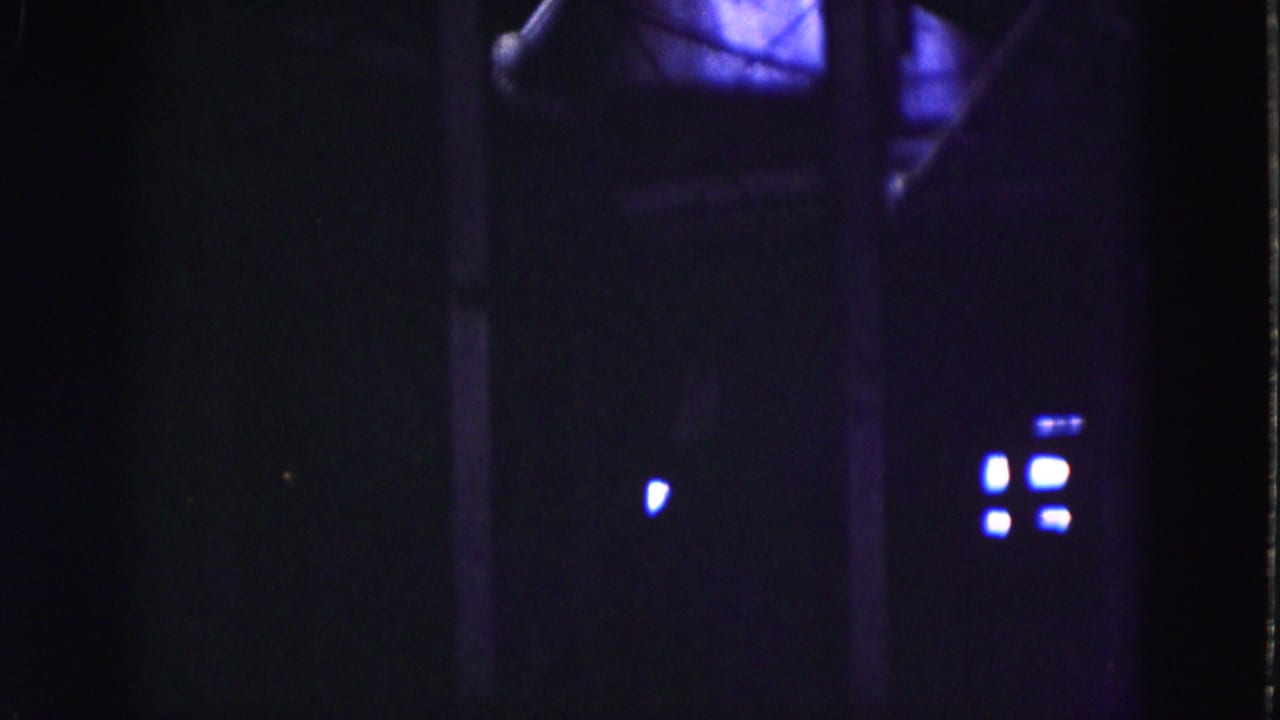 Low-light 1975 Everglades home-movie frame with a small bright light, purple-blue glow, and indistinct building forms.