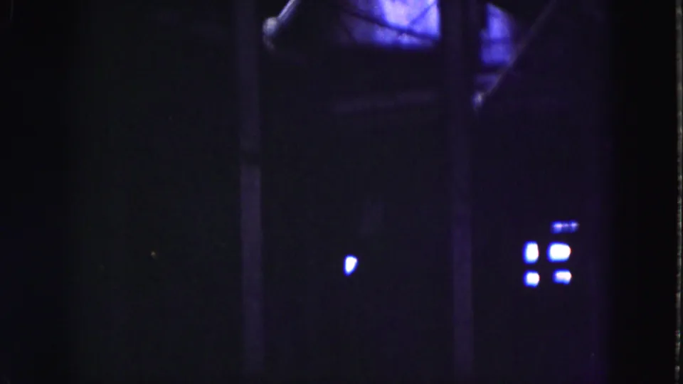 Low-light 1975 Everglades home-movie frame with a small bright light, purple-blue glow, and indistinct building forms.