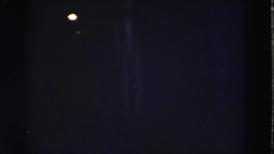 Grainy 8mm frame from the Florida Everglades clip showing a small warm light below dark vertical structural forms.