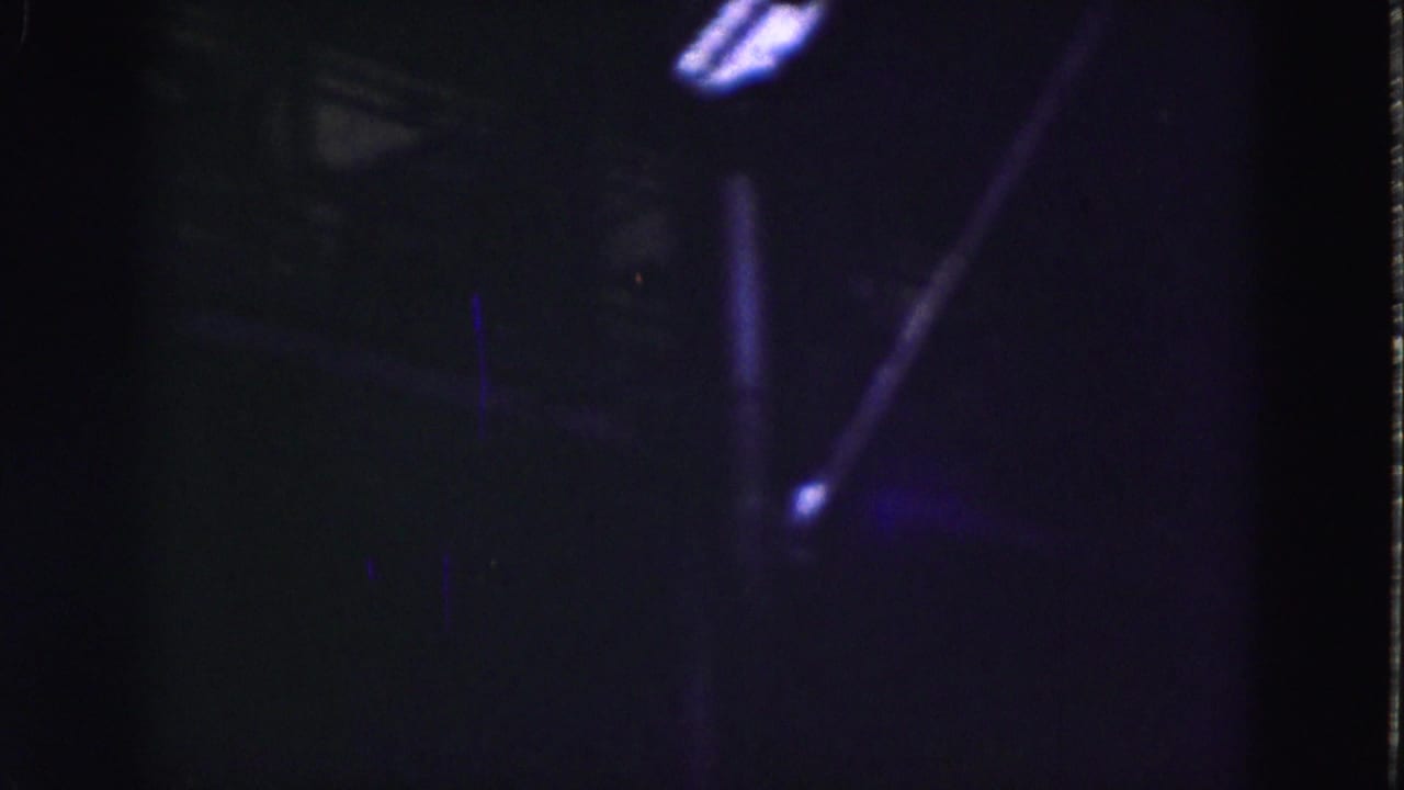Dim 1975 8mm view of an industrial interior in Florida, with structural beams and bright window openings in blue-purple light.