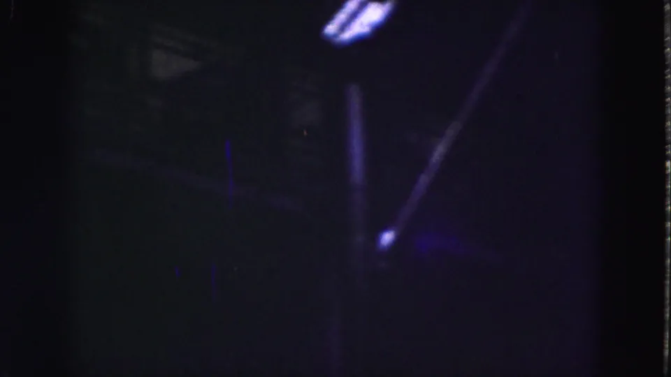 Dim 1975 8mm view of an industrial interior in Florida, with structural beams and bright window openings in blue-purple light.