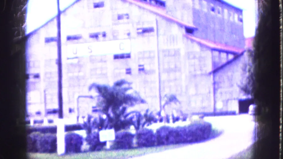 Exterior 1975 Florida view of a large brick industrial building with rows of windows, greenery, utility poles, and visible USC lettering.