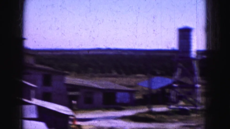 1975 Florida Everglades roadside scene showing a low building, raised tank, parked older cars, and open land in soft 8mm color.
