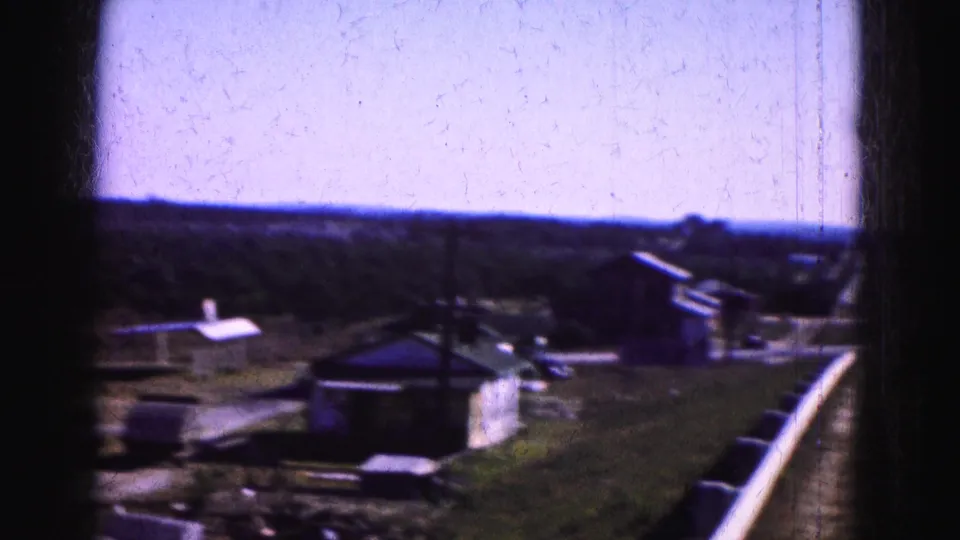 1975 Florida Everglades home-movie frame showing small roadside structures, open ground, distant trees, and a dark vertical film edge.