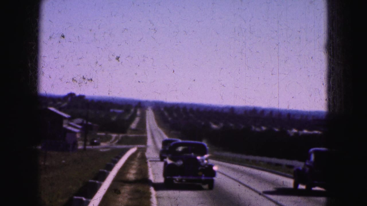Older cars travel along a straight road bordered by low buildings, vegetation, and a roadside barrier.