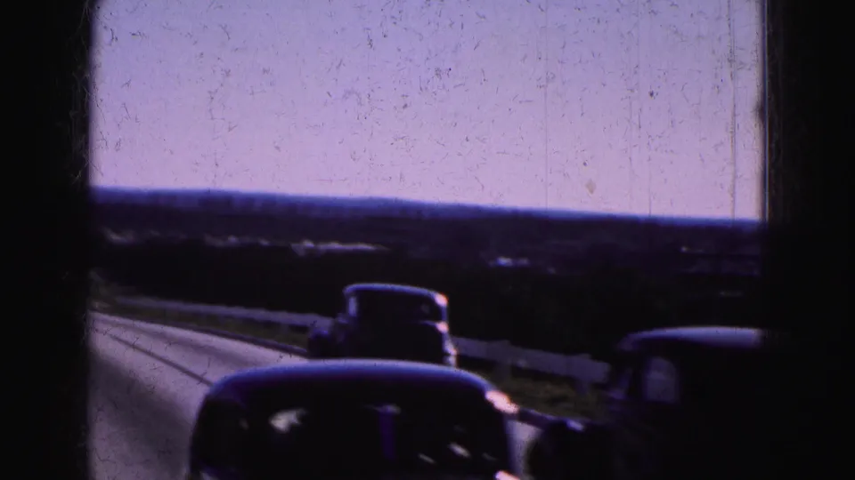 Close passenger-view 8mm frame from a 1975 Everglades road trip, with older cars passing on a rural highway under a pale sky.