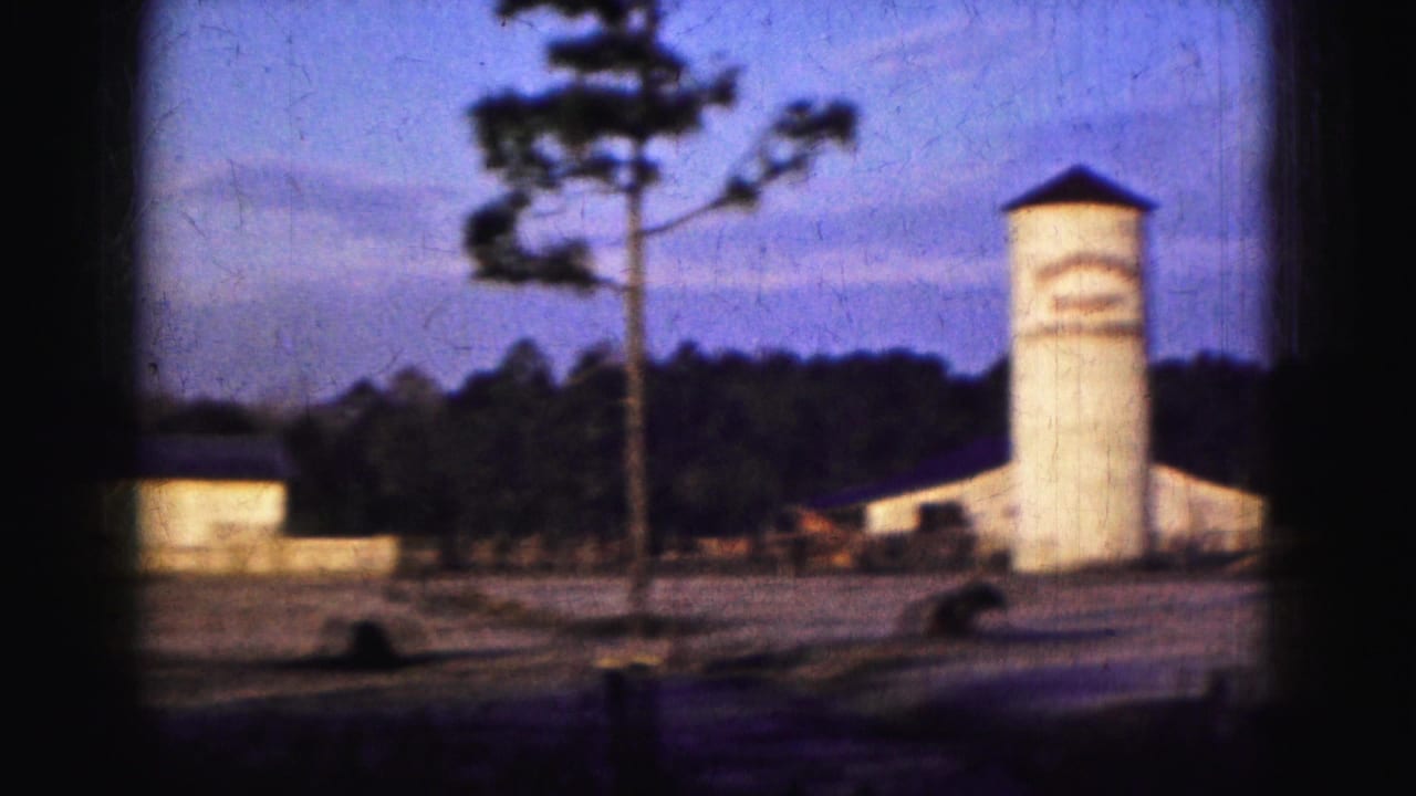 1975 Florida home-movie frame showing a white tower, low buildings, a pine tree, and a dark treeline under a blue dusk sky.