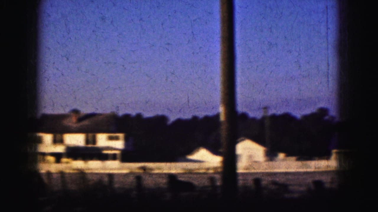 1975 Florida home-movie view of a low white building, vertical pole, small roofline, and dark trees in soft dusk light.