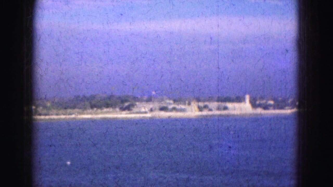1975 Florida waterfront frame showing calm blue water, a distant low strip of land, small shoreline structures, and a pale evening sky.