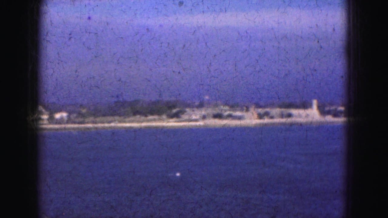 1975 Florida Everglades home-movie frame showing blue water, hazy sky, and a distant low shoreline with small structures.