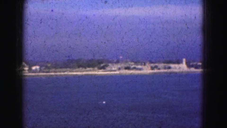 1975 Florida Everglades home-movie frame showing blue water, hazy sky, and a distant low shoreline with small structures.