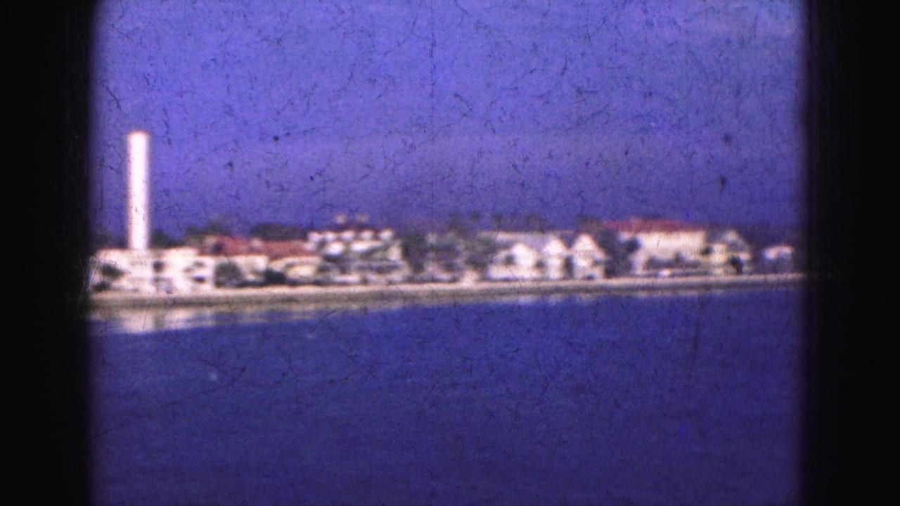 1975 8mm Florida waterfront view with a tall white tower and low buildings beyond calm blue water.
