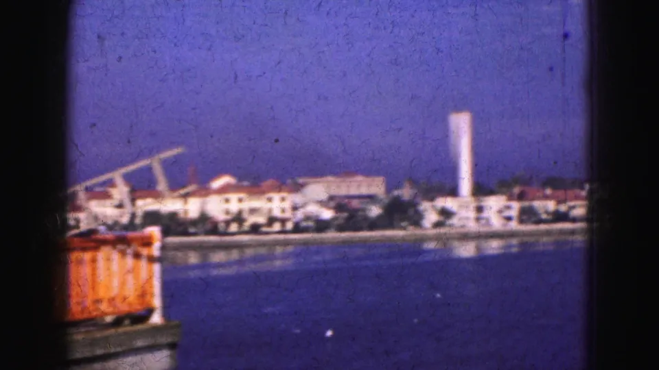 1975 8mm Florida shoreline view with white buildings, a tall tower, and a red-orange dockside object in the foreground.