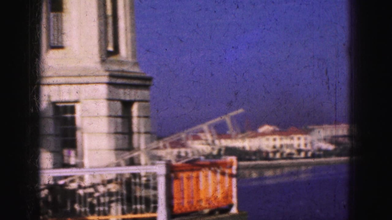1975 8mm Florida waterfront view with a building facade and railing foregrounding the distant shoreline across blue water.