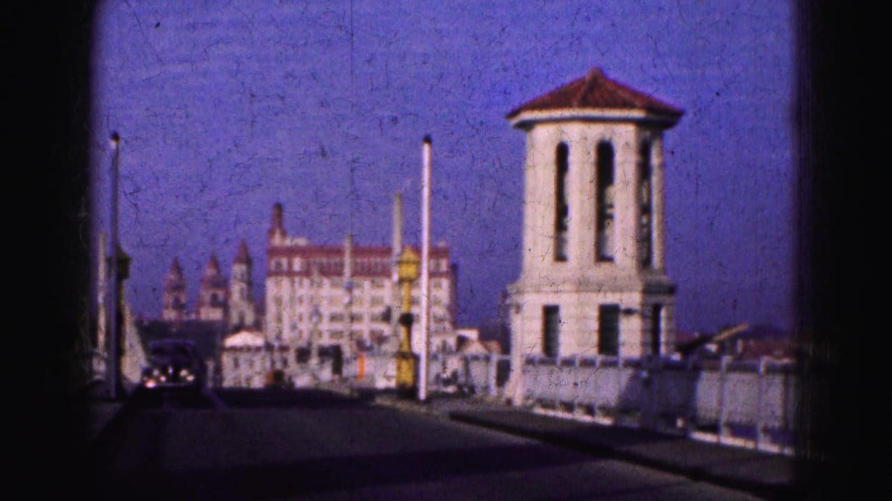 A 1970s 8mm view of a Florida roadway or bridge approach, with pale towers, cars, poles, and a large distant building.