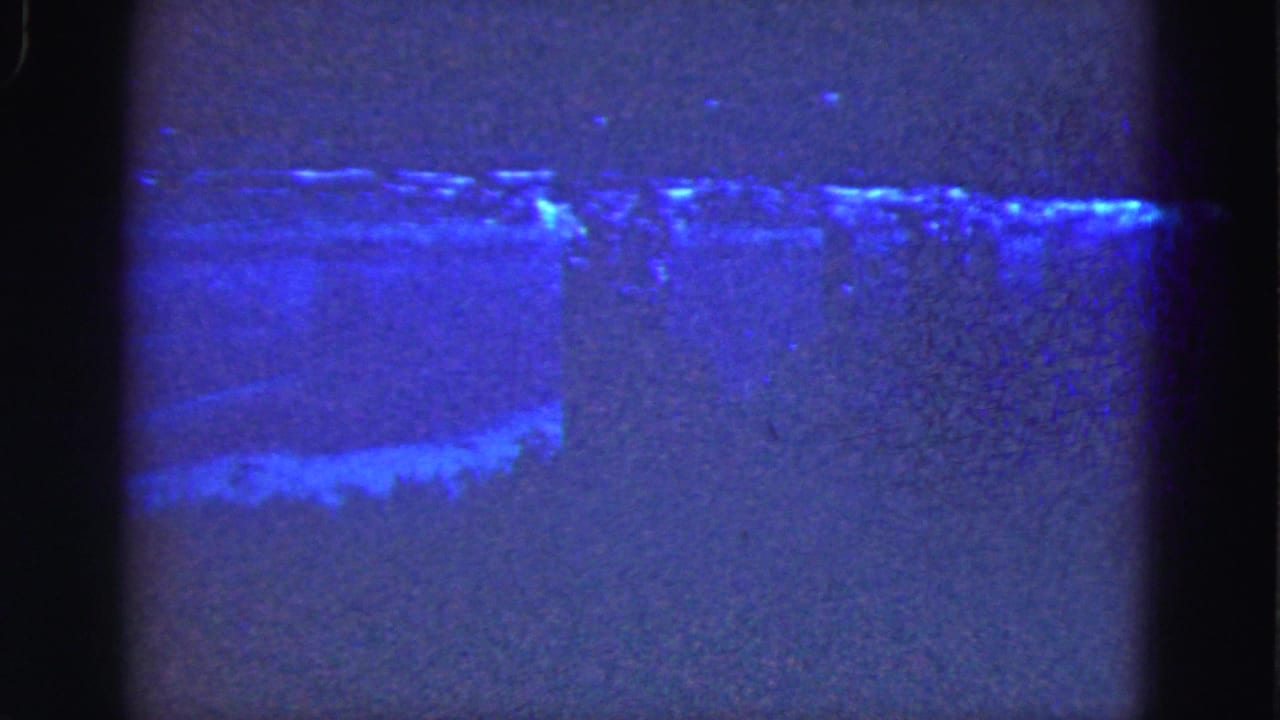 1975 Florida Everglades 8mm frame showing a dark blue shoreline or water edge with faint light patterns and visible film gate edges.