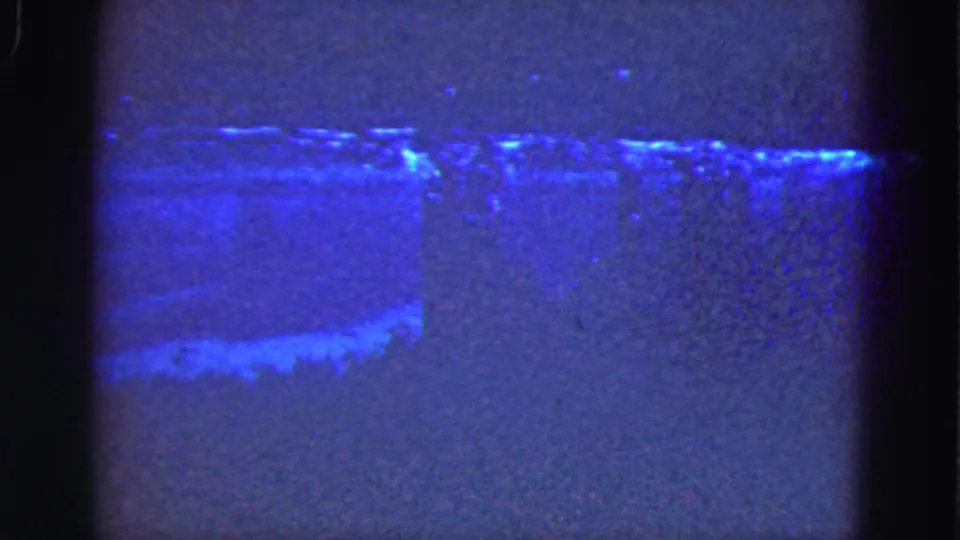 1975 Florida Everglades 8mm frame showing a dark blue shoreline or water edge with faint light patterns and visible film gate edges.