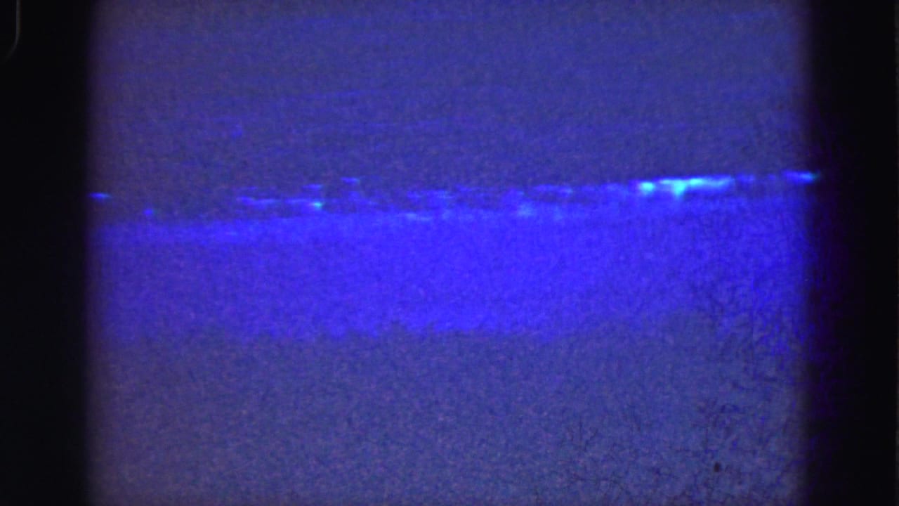 1975 Florida Everglades home-movie frame showing blue water, a low shoreline, and rounded dark film-gate edges.