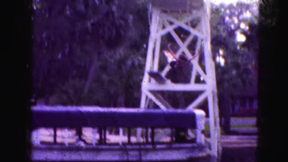 1975 Florida Everglades home-movie frame showing a white wooden lookout tower by a canal, with a small figure positioned near the structure.