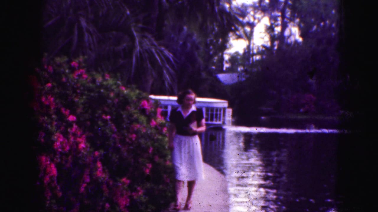 1975 Florida Everglades home-movie frame with a woman in a dark top and light skirt on a path beside bright flowering bushes and a canal.