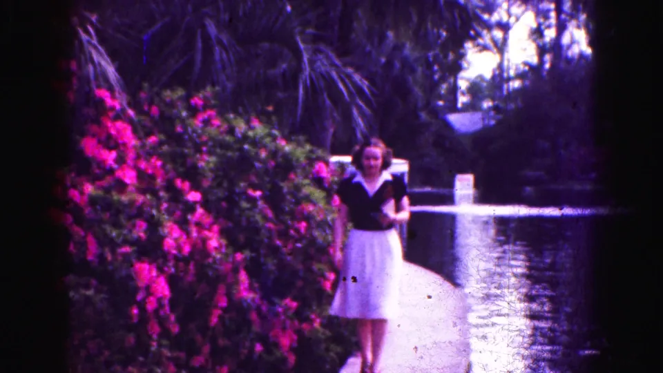 1975 Florida Everglades home-movie frame showing a woman walking beside a canal, with pink flowering bushes and trees filling the background.