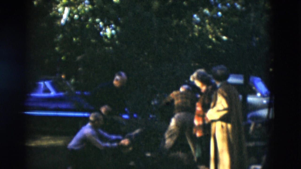 1975 Somerset, Kentucky home-movie frame showing adults near a car in a shaded wooded area, with one person crouched low.