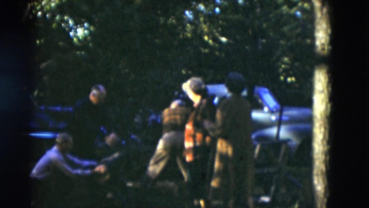 Adults in casual clothing stand and crouch near a parked car in wooded Somerset, Kentucky home-movie footage from 1975.