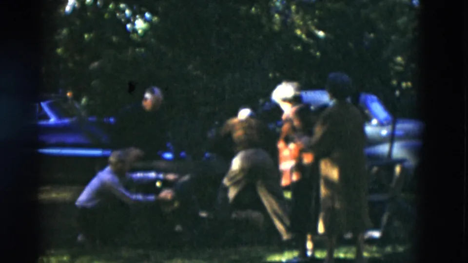 A 1975 home-movie still shows adults near a car under trees, including one person bending forward in the foreground.